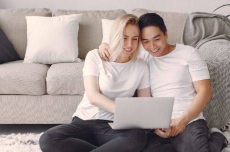 What You Need to Know Before Refinancing Your Home shows a couple sitting on the floor against a sofa while looking at a laptop.