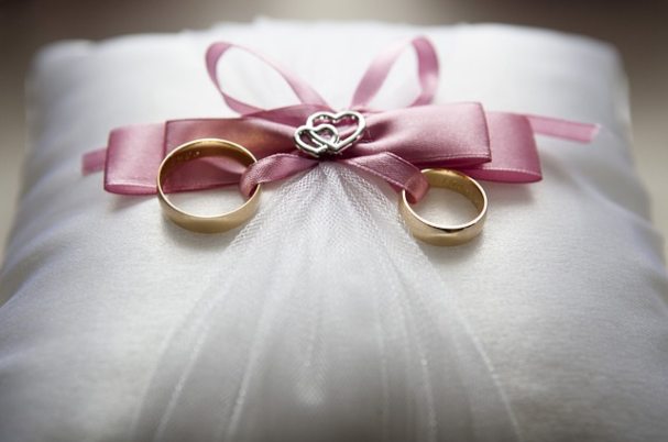 Spousal Debt image of wedding rings tied with pink ribbon to pillow.