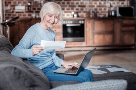 Social Security Claiming Strategies: A Guide for Widows and Widowers depicts elderly woman holding laptop and social security check.