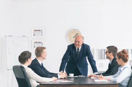 Bright office with formally-dressed managers gathered around a large table, with their smiling boss speaking about a successful family transfer of the business.