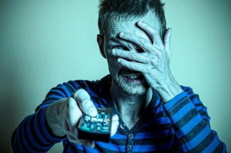 Elderly man covering eyes in rresponse to the stock market tumble while aiming remote control at TV