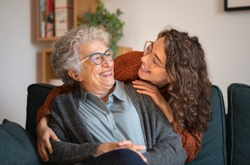 Assistance to Loved Ones photo concept with Old grandmother and adult granddaughter hugging at home and looking at each other. Happy senior mother and young daughter embracing with love on sofa. Happy young woman hugging from behind grandma with love.