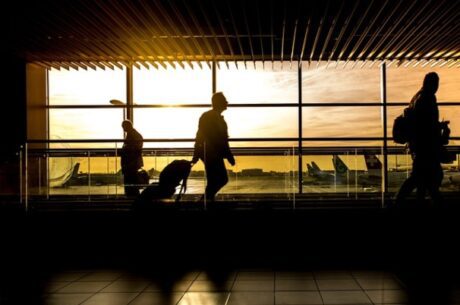 Do You Need Travel Health Insurance concept image depicts man pulling luggage through airport with pretty orange sunset in the window behind his silhouette.