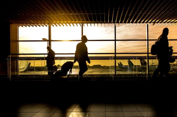Do You Need Travel Health Insurance concept image depicts man pulling luggage through airport with pretty orange sunset in the window behind his silhouette.