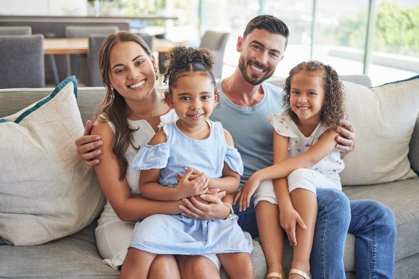 What You Should Know About Trusts concept image depicts a young husband, wife, and two young daughters sitting together on a white sofa.