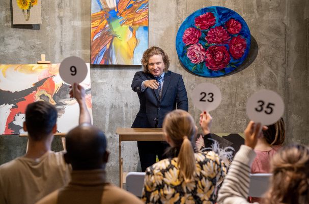 Should You Invest in Fine Art is depicted with a male auctioneer conducting an art auction with potential buyers holding up numbered paddles.