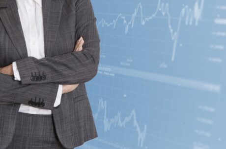 Quarter end report concept photo depicted by a business woman in a suit with arms crossed and standing in front of a large stock market screen.