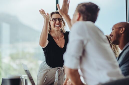 Employee Financial Wellness Program image depicts Businesswoman giving a high five to male colleague in meeting. Business professionals high five during a meeting in boardroom.