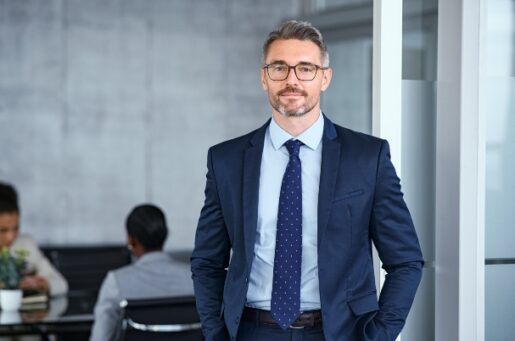 Financial Planning for Executives displays a Portrait of handsome mid adult business man standing in modern office. Successful mature entrepreneur in formal clothing looking at camera with satisfaction. Confident man in suit with eyeglasses and beard standing with hands in pocket and looking at camera in office.